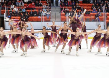 The front row of Haydenettes skaters skate toward the camera. Behind them, two lifts are taking place. The athletes wear dresses with purple tops and gold detail while the skirt has a mix of purple, yellow and orange.