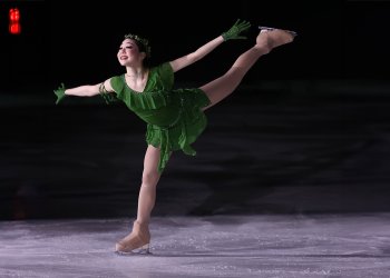 Koko Hirano performs a spiral on the ice. She is a young Asian woman wearing a green skating costume with a green leaf headband. 