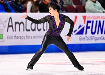 Tomoki Hiwatashi skates looking to the right with his right arm extended. He is a young man with short black hair wearing a black long sleeve top with a deep v neck and purple and blue beading around the collar. 