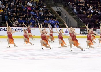 Photo of the Ice Denettes Affinity intermediate team. All athletes are in a coordinated kneeling position with their arms extended and smiles on their faces. They are all wearing matching skating costumes with a red jeweled top and orange skirt. 