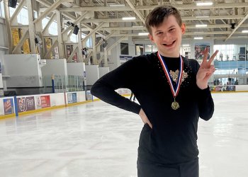 Tim poses on the ice with a medal around his neck. Tim is a white teenager with short brown hair. He is wearing a black long sleeved shirt with jewels on the chest. He stands with his right hand on his hip and his left hand making a peace sign