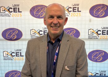 Photo of U.S. Figure Skating CEO Matt Farrell. Matt is an older white man wearing a navy blue button down shirt and a grey blazer. He stands in front of a backdrop with the 2025 Excel National Festival logo and the Broadmoor logo on it