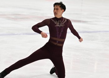 JY competes on the ice. They are an Asian skater with short hair wearing a maroon skating costume with jewels down the front. They are kneeling on their left knee with their right leg outstretched to the side