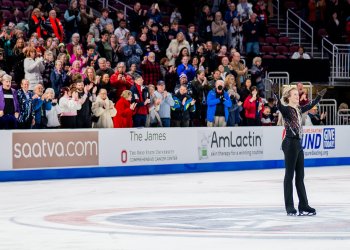 Ilia Malinin salutes the crowd with his arms outstretched and a smile on his face as people in the stands cheer. Ilia is a young man with short blonde hair wearing a black top with red and white stripes and black pants. 