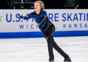 Ilia Malinin skates forward with his arms outstretched in front of him. He is a young man with short blonde hair wearing a dark blue long sleeve shirt with black stripes and black pants. 