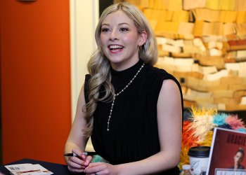 Gracie Gold at a book signing for her new book. She smiles, sitting at a table. She wears a black top.