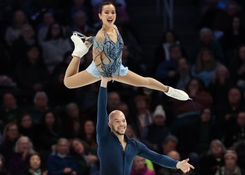 Ellie Kam and Danny O'Shea perform a lift with Danny holding Ellie up in the air, both smiling. Ellie is a young woman wearing a light blue skating costume and Danny is a man who is bald wearing a dark teal long sleeve top