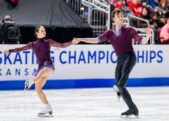 Katie McBeath and Daniil Parkman skate holding hands with one leg kicked out. Katie is a young woman with long black hair tied back in a bun wearing a long sleeve maroon skating costume. Daniil is a man with short brown hair wearing a coordinating maroon top and black pants