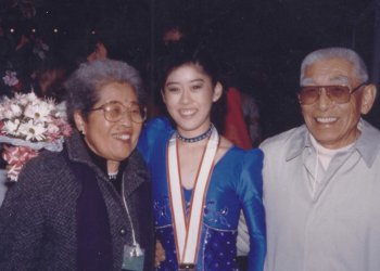Kristi poses for a photo with her grandparents. Kristi (center) is an Asian woman wearing a blue skating costume with puffy sleeves and a medal around her neck. Her grandmother (left) is an older Asian woman with short grey hair and glasses. She is wearing a grey coat. Her grandfather (right) is an older Asian man with grey hair and glasses. He is wearing a white button up shirt 