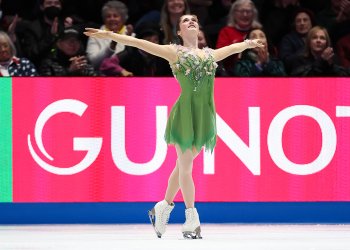 Isabeau Levito acknowledges the crowd with her arms outstretched and a big smile on her face. She is wearing a green dress with light pink floral details at the shoulders. She has brown hair that is tied back in braids. 
