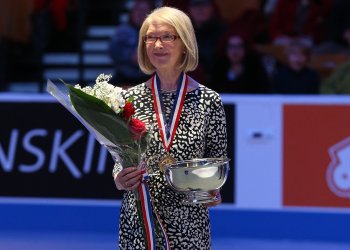 Lynn Benson stands at center ice, holding flowers and a ceremonial bowl, as she is inducted into the U.S. Figure Skating Hall of Fame.