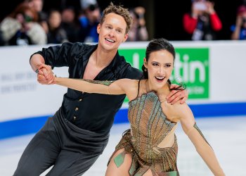 Madison Chock and Evan Bates skate hand in hand. Madison is a young woman with long black hair wearing a copper and green dress inspired by the statue of liberty and Evan is a young man with short curly blonde hair wearing a button down black shirt with the sleeves rolled up to the elbows. 
