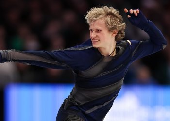 Dressed in all black, Ilia Malinin performs a routine. He is smiling and pointing straight out with one arm and has the other arm pointing up
