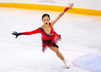Sherry Zhang competes at JGP Austria. She is a young woman with black hair tied back in a low bun. She is wearing a long sleeve red skating costume. Shr skates on one leg with her arms outstretched