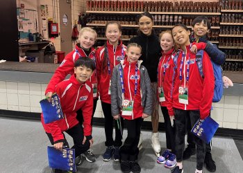 Molli Taylorson and Ethan Peng along with five other young skaters pose for a photo with Team USA's Madison Chock. Ethan is a young Asian boy with braces. Molli is a young white girl with blonde hair tied back into braids. All of the young skaters are wearing red jackets with the U.S. Figure Skating log on the front and the words National Development Team on the back. 