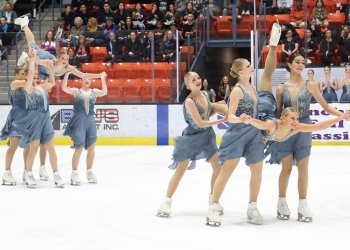 The Northernettes, in clue dresses with flowy skirts and beaded tops, perform two lifts - one in the forefront and one in the background. In both, three athletes are holding a girl on her stomach as she floats down toward the ice, her right leg up in the air.