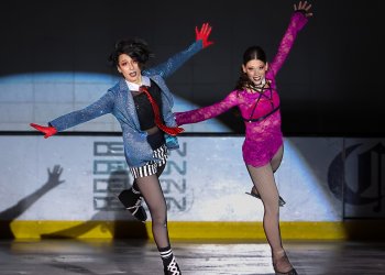 Ashlee Pantano (left) and Ashlyn Olson (right) skate side by side with their arms extended outward. Ashlee is wearing a black collared shirt with a red tie and a sparkly blue blazer over top with black and white striped shorts. Ashlyn is wearing a hot pink top with a black strap over her chest. 