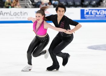 Yahli Pedersen, in a pink crop top and black leather pants, and Jeffrey Chen, wearing all black with pink detail diagonally down his shirt, smile while in hold during their rhythm dance.