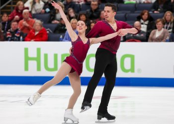 Valentina Plazas and Maximiliano Fernandez compete in the short program at the 2024 Prevagen U.S. Figure Skating Championships. Plazas (left) is a woman with black hair tied back in a tight bun. She is wearing a red skating costume. Fernandez (right) is a man with short black hair wearing a red long sleeve top and black pants. Plazas skates with her arms lifted above her head and her right leg extended outward. Fernandez stands behind her holding her hand and placing his other hand on her waist 