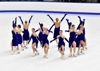 Hayden Select competes wearing matching dark blue skating costumes with a silver neckline. They all stand in a circle facing out and each skater is alternating with their arms up in the air or extended downward