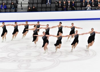 Hayden Select skates in two lines with each skater holding each other by the shoulder. They are wearing matching one shoulder black skating costumes