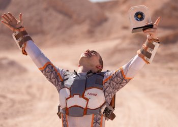 Adam Rippon holds his hands up in the air looking victorious. He is wearing a suit resembling a space suit for Stars on Mars and is holding a trophy in his right hand
