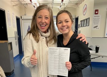 Sandy Lenz (left) poses for a photo from the waist up with her adult student, Emily Thompspon (right). Lenz is an older white woman with shoulder length blonde hair. She is wearing a white sweater with a white scarf and gives the camera a thumbs up with her other arm around the shoulder of her student. Emily is wearing a black long sleeve skating costume and it proudly holding up her testing papers with a big smile. 