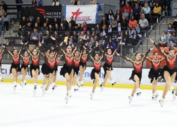 Skyliners junior, in dresses with a black shirt and fire going up the bodice, all jump at the same time toward the camera while on the ice. Behind them in the background is a crowd. 
