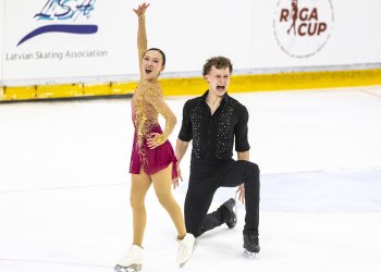 Pairs Naomi Williams and Lachlan Lewer strike a dramatic ending pose at JGP Latvia. Williams, wearing a red dress, has one arm on her hip and one extended into the air. Lewer, dressed in black, is on a knee with mouth wide open. 