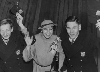 old black and white photo of two men and a woman smiling and waving at the camera. The two men are wearing black coats with the U.S. Figure Skating emblem on the front. The woman stands between them wearing a long coat with fkowers pinned to the front. She has short curly hair. 