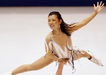 Kyoko Ina is held up above her partners head in a lift while performing at the 2002 Winter Olympic Games in Salt Lake City. Kyoko is a young Asian woman with long black hair. She is wearing a white skating costume and white skates. 