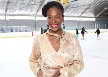 Kadari Taylor-Watson poses on the ice with her Trailblazer Award. She is a Black woman with her hair up. She is wearing large black curcular earrings and a gold top with a bow around the neck. She is holding a glass award which is in the shape of a tear drop