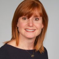 Headshot of a Caucasian woman smiling. She has shoulder length red hair with bangs. She is wearing a black shirt and a dainty necklace. 