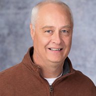 Headshot og \\f Troy Schwindt. He is an older Caucasian man with grey hair. He is wearing a burnt orange quarter-zip jacket with a white tshirt on underneath