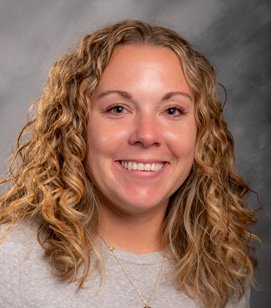Headshot of a Caucasian woman smiling. She has curly blonde hair and is wearing a grey shirt. 