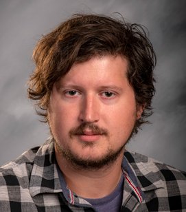 Headshot of a Caucasian man. He has brown hair and a mustache. He is wearing a black and grey plaid shirt