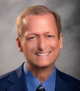 Headshot of a Caucasian man smiling. He has short blonde hair and is wearing a light blue collared shirt and black blazer