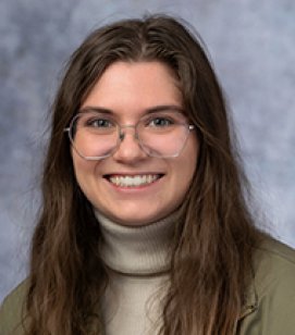 Headshot of Meg Metzger-Seymour. She is a Caucasian woman with long brown hair. She is wearing a grey turtleneck with a green jacket on top and glasses
