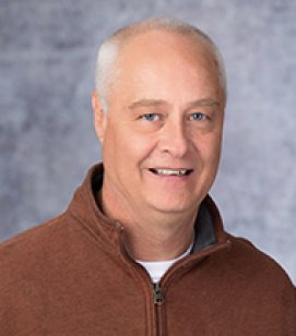 Headshot og \\f Troy Schwindt. He is an older Caucasian man with grey hair. He is wearing a burnt orange quarter-zip jacket with a white tshirt on underneath
